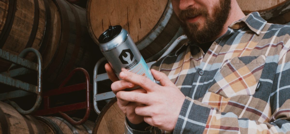 Moersleutel brewer reading a beer can label in front of stacked wooden barrels in the brewery