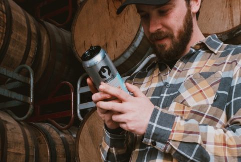 Moersleutel brewer reading a beer can label in front of stacked wooden barrels in the brewery