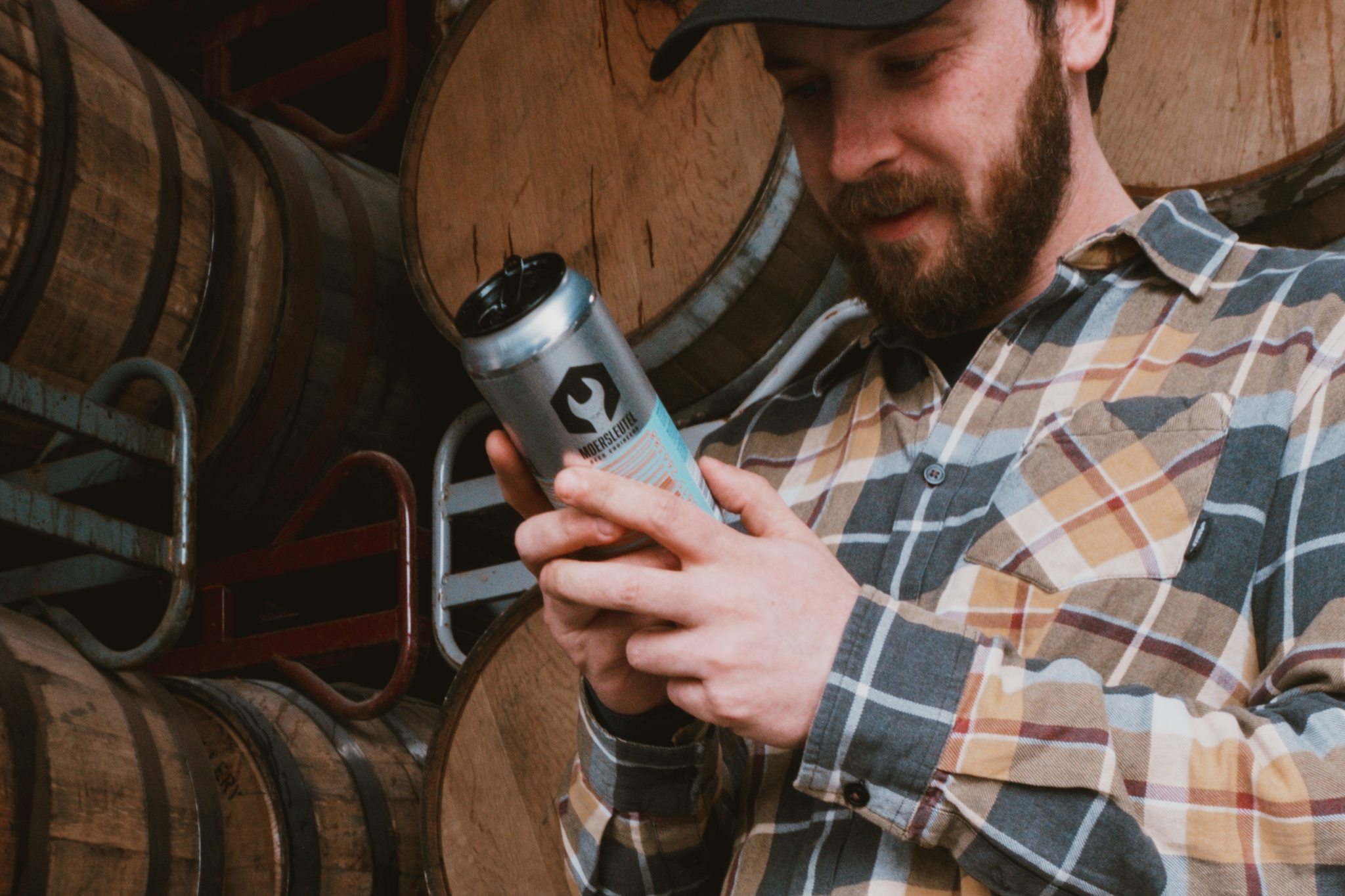 Moersleutel brewer reading a beer can label in front of stacked wooden barrels in the brewery