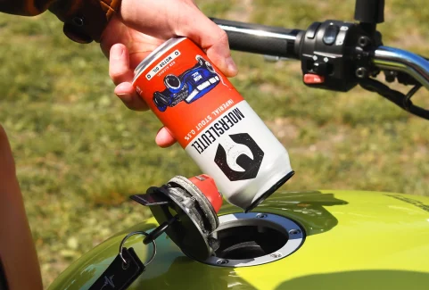 Person playfully pouring Moersleutel Motor Oil Non-Alcoholic Stout into a motorcycle fuel tank